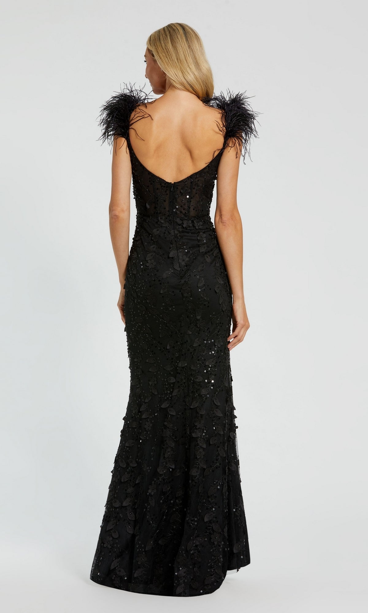A woman wearing the Long Formal Dress 20889 by Mac Duggal—black, floor-length with sequin embellishments, feathered straps, dramatic v-neckline, and elegant feathered shoulder details—stands facing away from the camera.